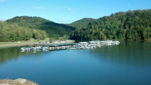 A serene lake surrounded by green hills, featuring a marina with several boats docked. Clear blue sky above.
