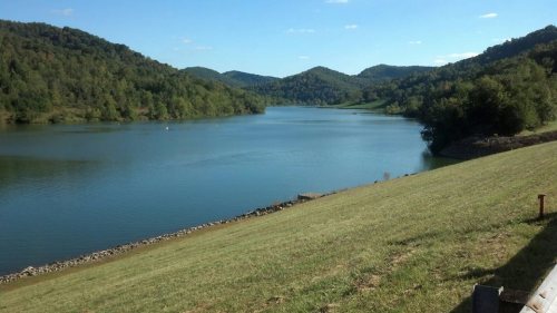 A serene lake surrounded by green hills under a clear blue sky, with gentle slopes leading to the water's edge.