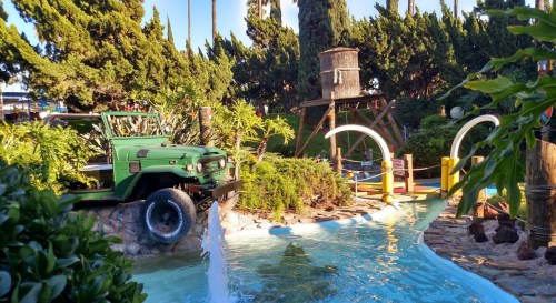 A vibrant mini-golf scene featuring a green jeep, lush greenery, and playful water features under a clear blue sky.