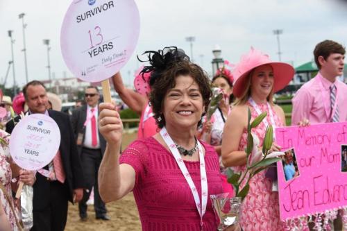 A smiling woman in a pink dress holds a sign reading "SURVIVOR 13 YEARS" at a pink-themed event with others in the background.