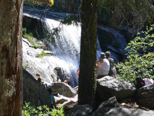 A serene waterfall surrounded by rocks and trees, with people sitting nearby enjoying the natural scenery.