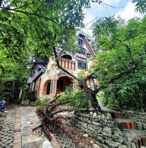 A colorful, multi-story house surrounded by lush greenery and trees, with a stone pathway leading to the entrance.