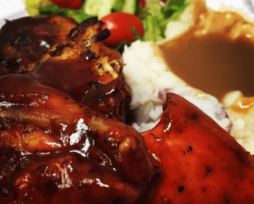 A close-up of grilled chicken with barbecue sauce, served with mashed potatoes, gravy, and a side salad with tomatoes.