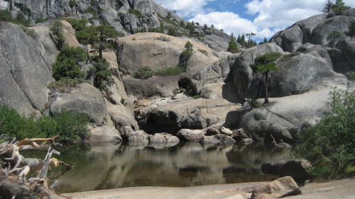 A serene landscape featuring large rocks and a calm pool of water surrounded by greenery under a blue sky.
