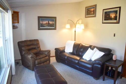 Cozy living room with a leather sofa, armchair, ottoman, and framed artwork on the walls. Soft lighting from a floor lamp.