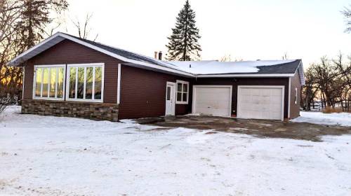 A brown house with large windows and a stone foundation, surrounded by snow and trees, featuring a double garage.