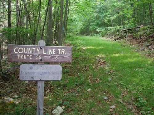 A wooden sign marks the start of County Line Trail, with a grassy path leading into a wooded area.