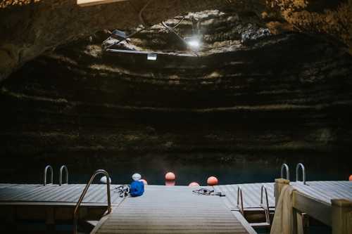 A wooden dock leads into a dark cave with red buoys floating in the water, illuminated by overhead lights.