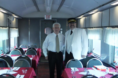 Two men in formal attire stand inside a dining car, surrounded by tables set for a meal.