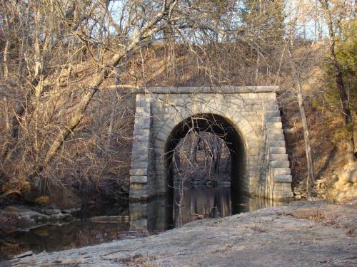 Stone arch bridge over a calm stream, surrounded by bare trees and rocky terrain in a serene natural setting.