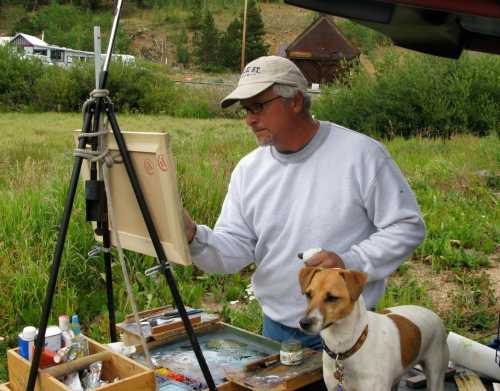 An artist paints on an easel outdoors, with a dog sitting beside him in a grassy area.