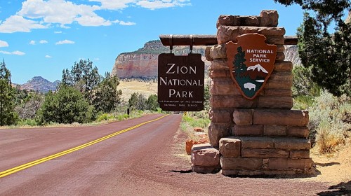 Sign for Zion National Park at the entrance, with a scenic landscape and blue sky in the background.