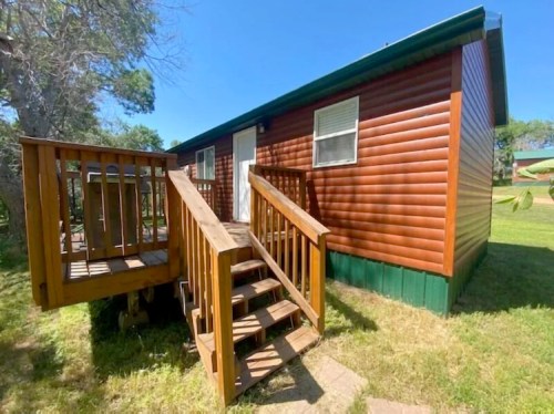 A wooden cabin with a sloped roof, featuring a deck and stairs, surrounded by green grass and trees.
