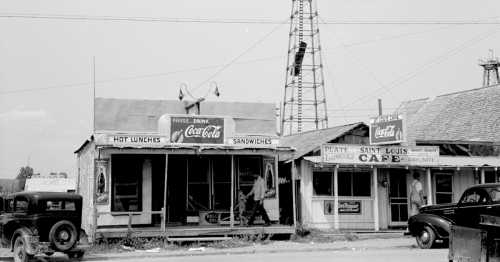 Black and white photo of vintage storefronts, featuring signs for Coca-Cola and a café, with a car parked nearby.