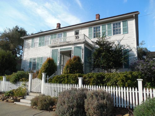 A large white house with green shutters, a balcony, and a white picket fence, surrounded by greenery.