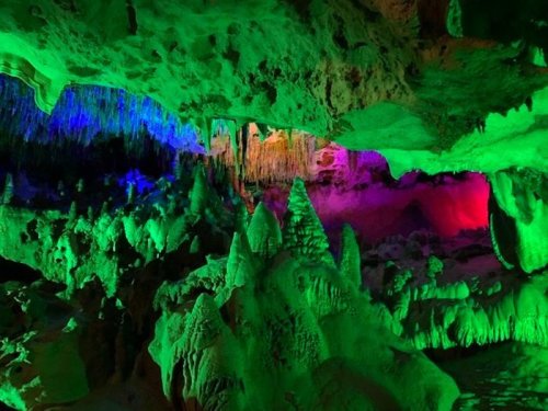 A vibrant cave interior illuminated with green, purple, and blue lights, showcasing unique rock formations and stalactites.