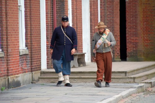 Two men in historical costumes walk along a brick pathway, one in a military uniform and the other in casual attire.