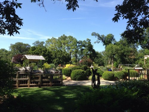 A sunny garden scene with lush greenery, neatly trimmed bushes, and a wooden gazebo under a clear blue sky.