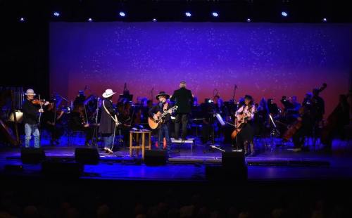 A musical performance on stage with musicians, an orchestra, and colorful lighting in the background.