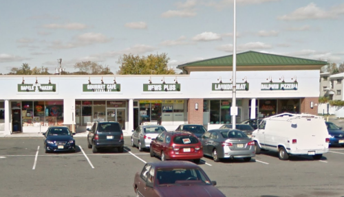 A shopping plaza with various storefronts, including a bakery, café, and pizzeria, surrounded by parked cars.