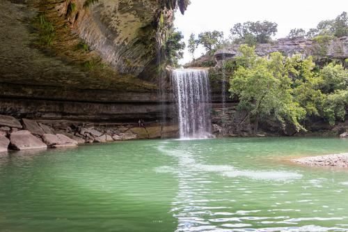 A serene waterfall cascades into a green pool, surrounded by rocky cliffs and lush trees.