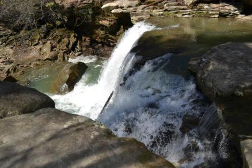 A waterfall cascading over rocks into a clear pool, surrounded by lush greenery and rugged terrain.