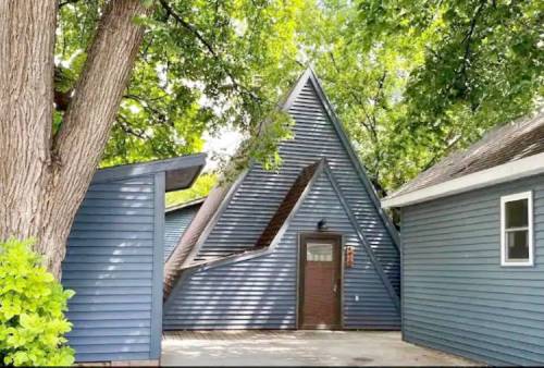 A unique triangular-shaped house surrounded by trees and two adjacent buildings.