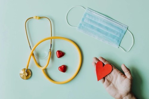 A hand holds a red paper heart next to a stethoscope, two chocolate hearts, and a blue medical mask on a light background.