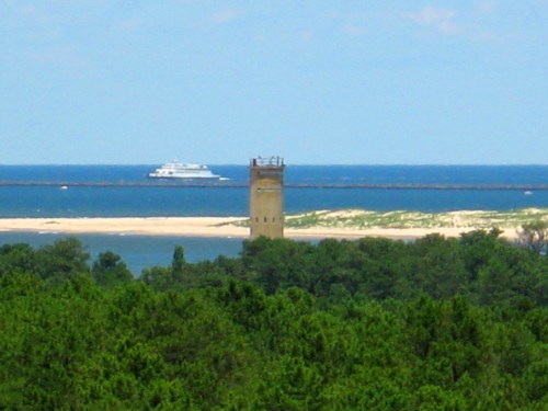 A coastal scene featuring a tower surrounded by greenery, with a boat sailing on the water in the background.