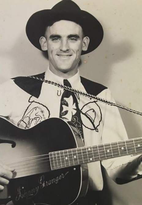 A smiling man in a cowboy hat holds a guitar, wearing a decorated shirt with "Johnny Granger" written on it.