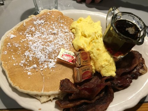 A plate with a large pancake topped with powdered sugar, scrambled eggs, bacon, and a small pitcher of syrup.