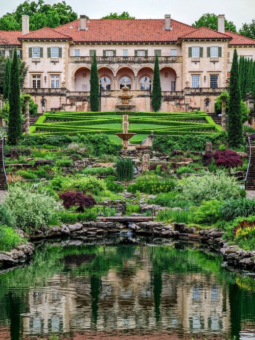A grand mansion with a red-tiled roof surrounded by lush gardens and a reflecting pond in the foreground.