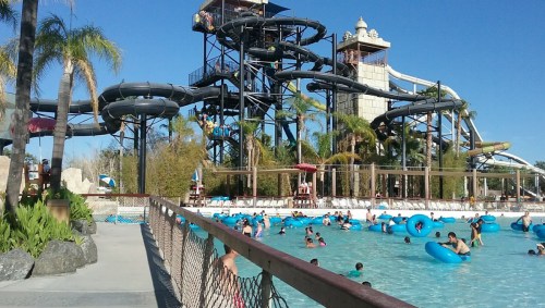 A water park scene with a large slide, palm trees, and people floating in a pool on inner tubes.