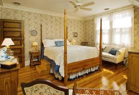Cozy bedroom with a four-poster bed, floral wallpaper, wooden furniture, and a comfortable seating area.