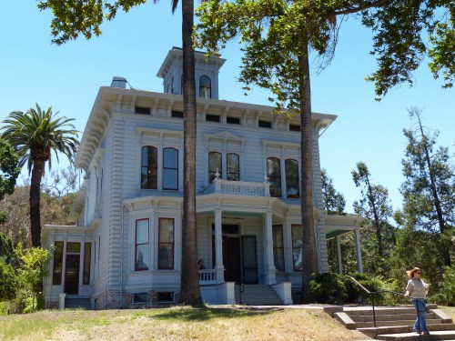 A large, historic blue house surrounded by palm trees and greenery, with a person standing on the steps.
