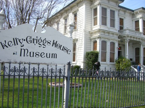 A historic white house with a sign reading "Kelly-Griggs House Museum," surrounded by a green lawn and fence.