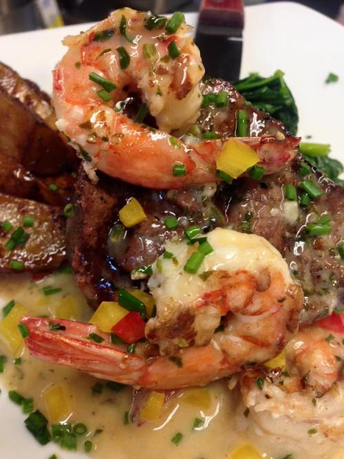 A plate of shrimp and steak topped with sauce, garnished with chives and colorful peppers, served with roasted potatoes.