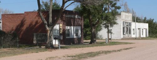 Abandoned buildings line a dirt road in a small, rural town, with trees and clear blue sky in the background.