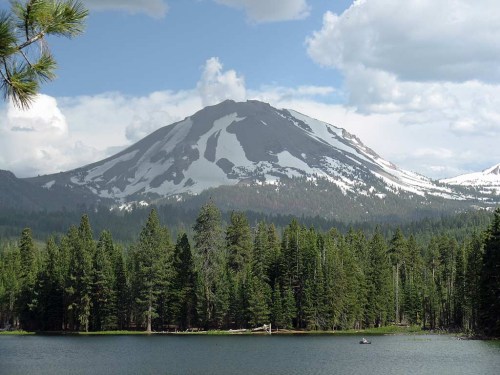 A scenic view of a snow-capped mountain surrounded by lush green forests and a calm lake under a partly cloudy sky.
