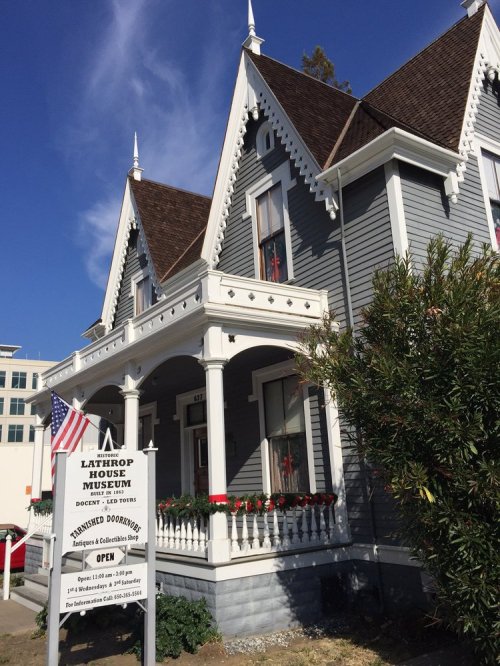 Victorian-style house museum with a sign, American flag, and decorative plants in front. Clear blue sky above.
