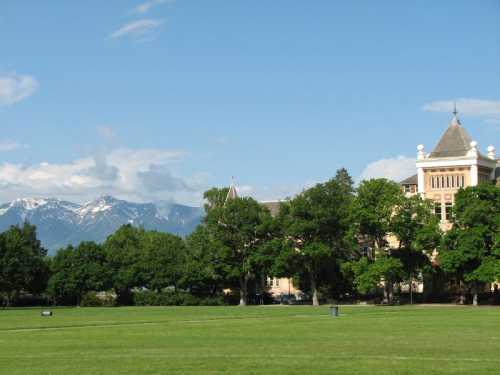 A grassy field with trees and a historic building, set against a backdrop of snow-capped mountains and a blue sky.