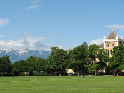 A grassy field with trees and a historic building, set against a backdrop of snow-capped mountains and a blue sky.