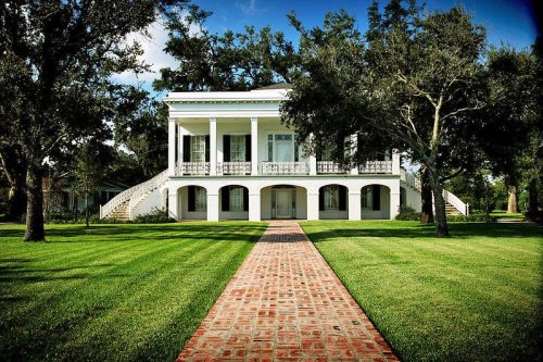 A grand white house with columns, surrounded by lush green grass and trees, with a brick pathway leading to the entrance.