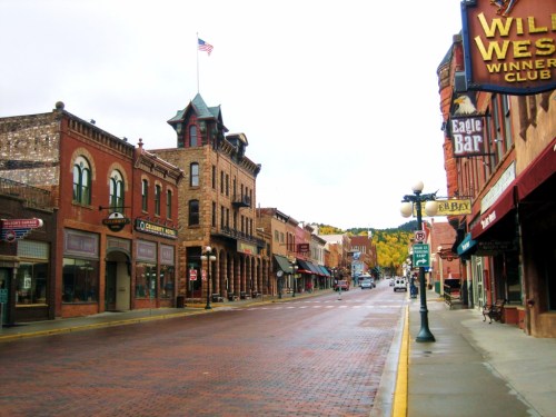A historic street lined with brick buildings, shops, and a flagpole, set against a backdrop of autumn trees.