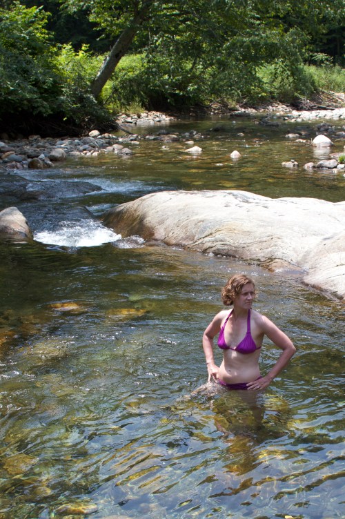 A woman in a purple bikini stands in a shallow stream, surrounded by rocks and greenery.