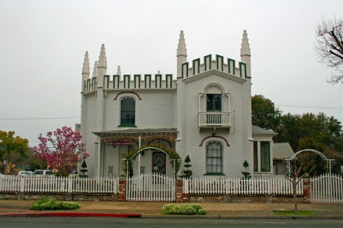 A whimsical, castle-like house with spires, a white picket fence, and colorful landscaping on a cloudy day.