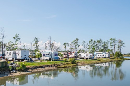 A row of RVs parked by a calm waterway, surrounded by trees and a clear blue sky.