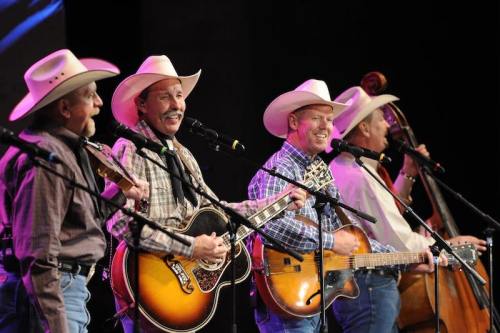 Four musicians in cowboy hats perform on stage with guitars and a double bass, smiling and singing into microphones.