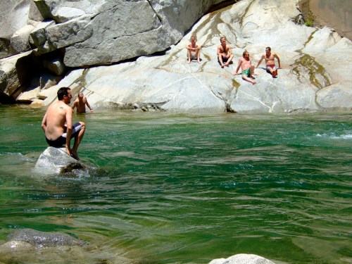 A group of people relax on rocks by a clear, green river, while one person sits on a rock in the water.
