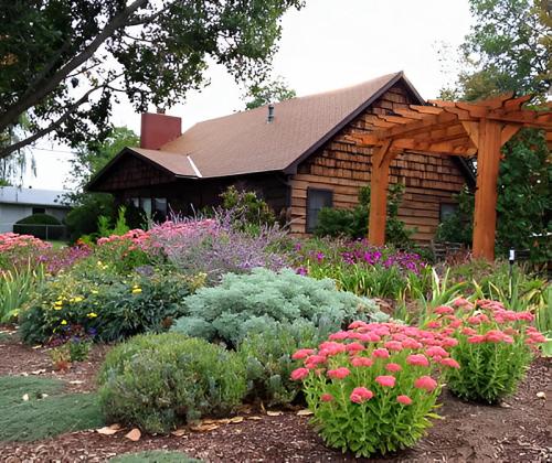 A charming wooden house surrounded by colorful flowers and lush greenery, with a wooden arbor in the garden.